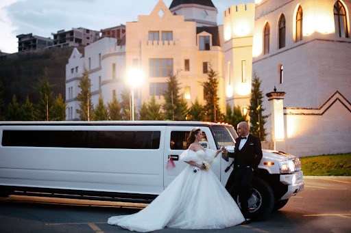 A bride and groom stepping out of a white stretch limousine outside their floodlit castle venue at dusk